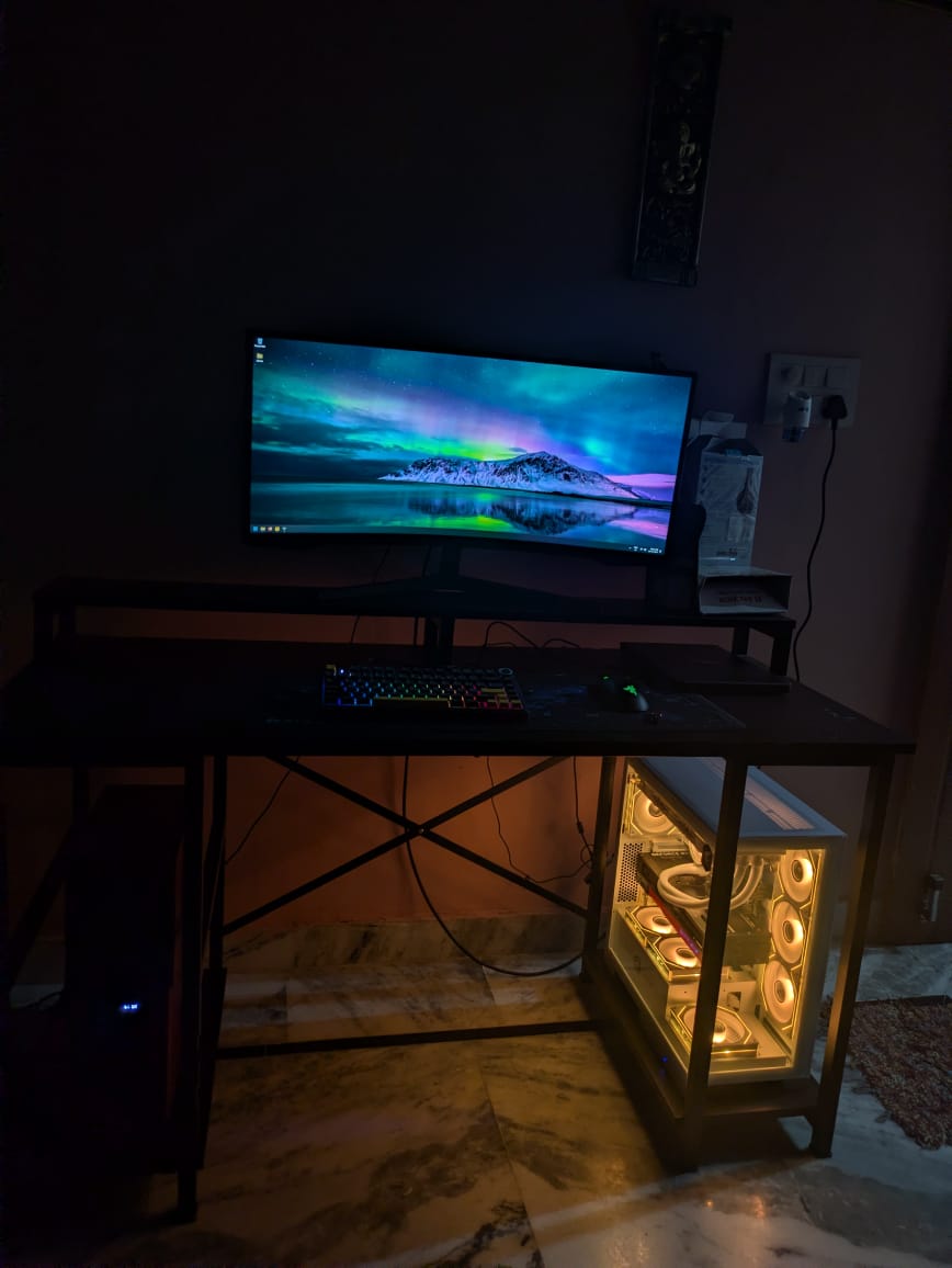 A dark room with a desk, an ultrawide monitor showing an aurora wallpaper, an RGB keyboard, and a white PC case glowing orange under the desk.